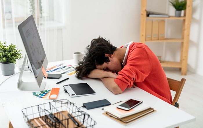 exhausted young male freelancer napping on desk in front of computer screen among working supplies and gadgets resize
