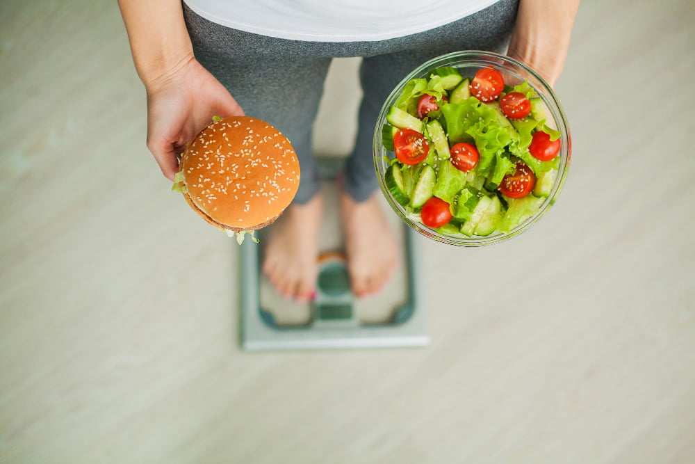 woman measuring body weight on weighing scale holding burger and salad sweets are unhealthy junk food dieting healthy eating lifestyle weight loss obesity top view