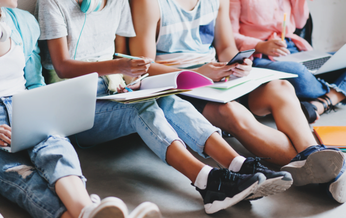 girl in blue jeans and black sneakers writing lecture in big textbook sitting on the floor with college friends young man typing message on phone while other students working with laptops